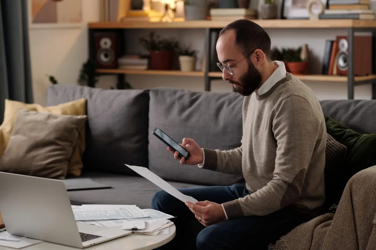 Homem sentado no sofá olha o celular enquanto segura um documento de papel, com laptop e contas sobre a mesa de centro