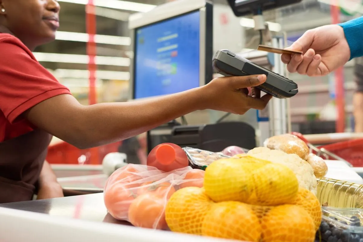 Cliente aproxima o cartão da maquininha segurada pela operadora de caixa, com frutas e pães na esteira do supermercado