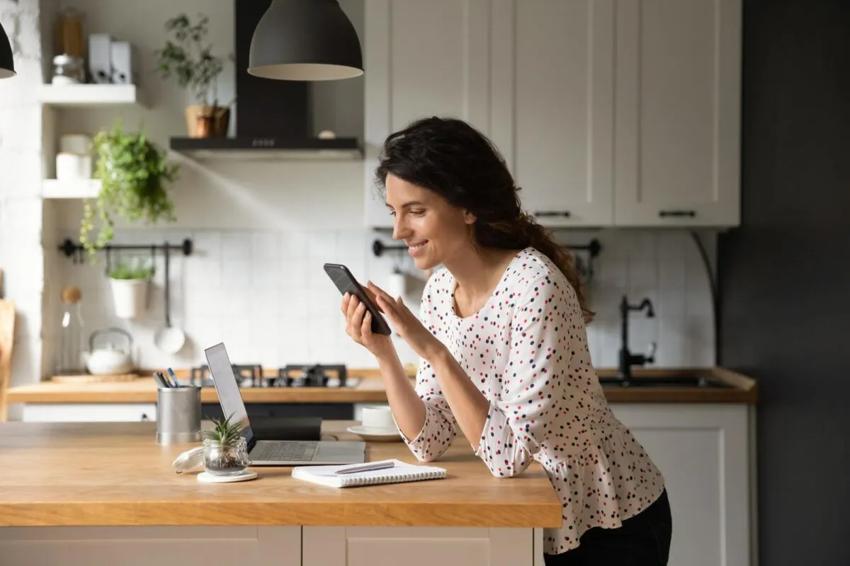 Mulher sorrindo usa o celular em pe numa cozinha moderna, com laptop e caderno sobre a bancada de madeira