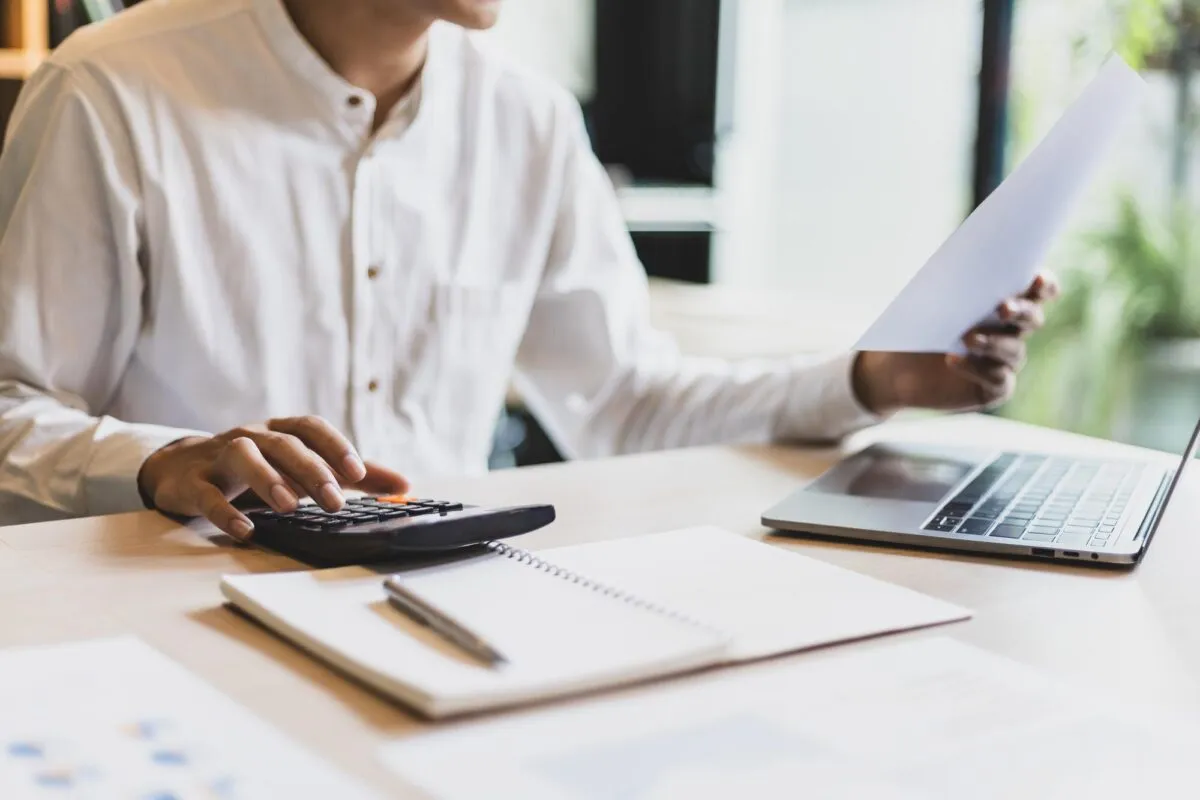 Homem usa calculadora enquanto revisa um documento financeiro sobre a mesa, ao lado de laptop e caderno