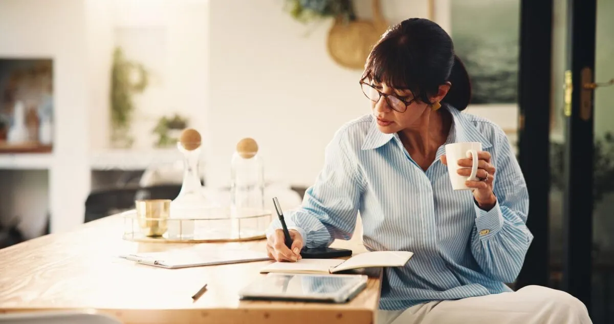 Mulher anota contas em um caderno enquanto toma café, organizando as finanças da empresa