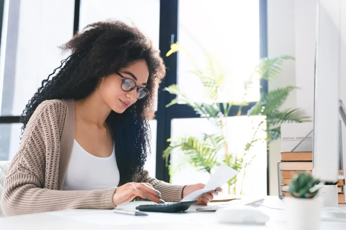 Mulher sentada à mesa usando calculadora e segurando um papel para organizar as contas do mês