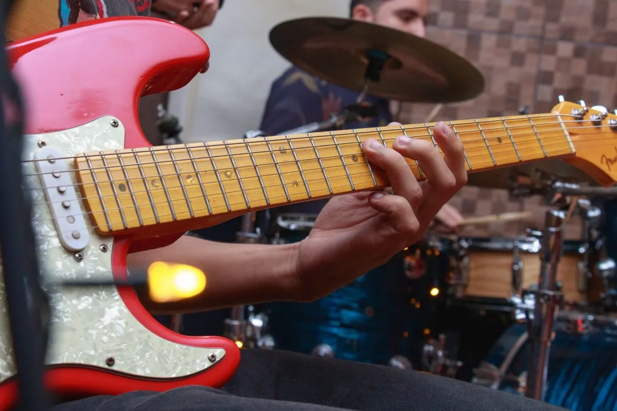 Músico de banda tocando guitarra elétrica em apresentação ao vivo, com baterista ao fundo no palco